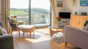 Ein modernes Wohnzimmer mit einer großen Schiebeglasdoor zum Balkon, einem Flachbildfernseher und einem Essbereich mit Tisch und Stühlen.