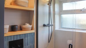 Une salle de bain moderne dotée d’une douche à paroi de verre, d’une étagère en bois avec des paniers et d’une fenêtre laissant entrer la lumière naturelle.
