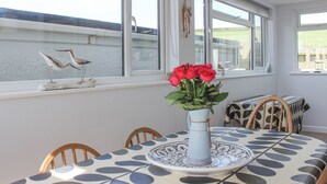 Un coin repas avec une table dressée pour deux personnes, un vase rempli de fleurs rouges et une vue sur la mer, visible à travers de grandes fenêtres.