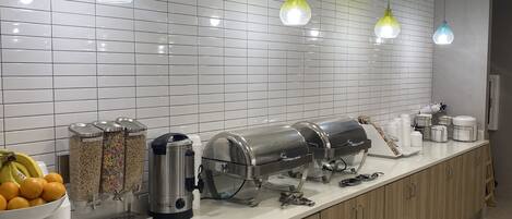 A modern kitchen with a long countertop, equipped with coffee machines, cereal dispensers, and a fruit bowl.