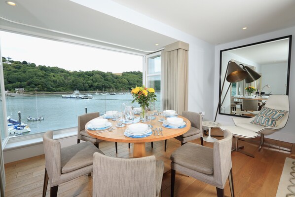 A dining area with a view of a harbor, a round table set with plates and cutlery, and a sofa with a mirror and cushions.