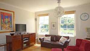 A living room with a brown leather sofa, a red armchair, a wooden cabinet with a television, and a chandelier. - Fairview House (Dartmouth)