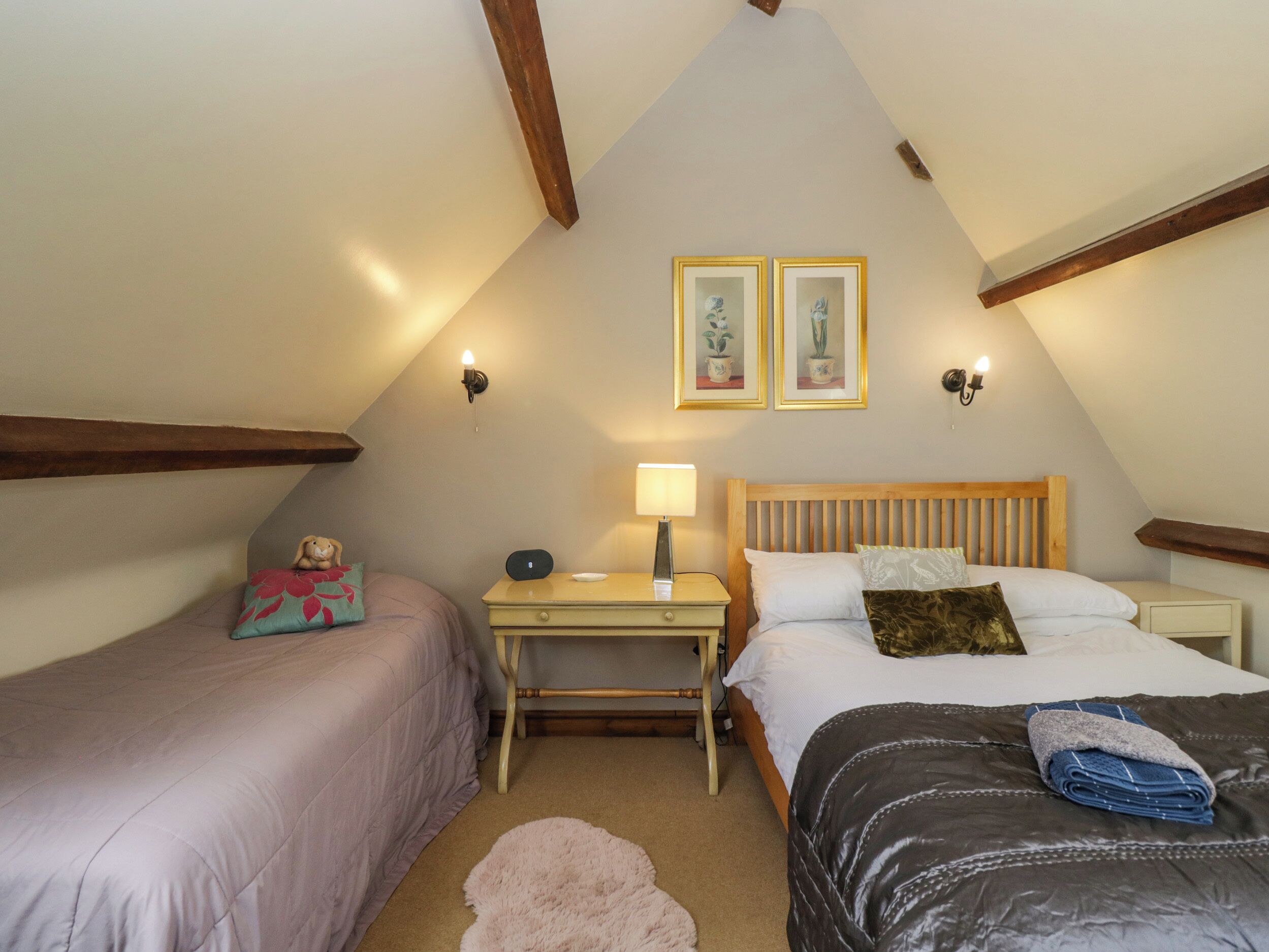 A cozy attic bedroom with two beds, a nightstand, a lamp, and two framed pictures on the wall.