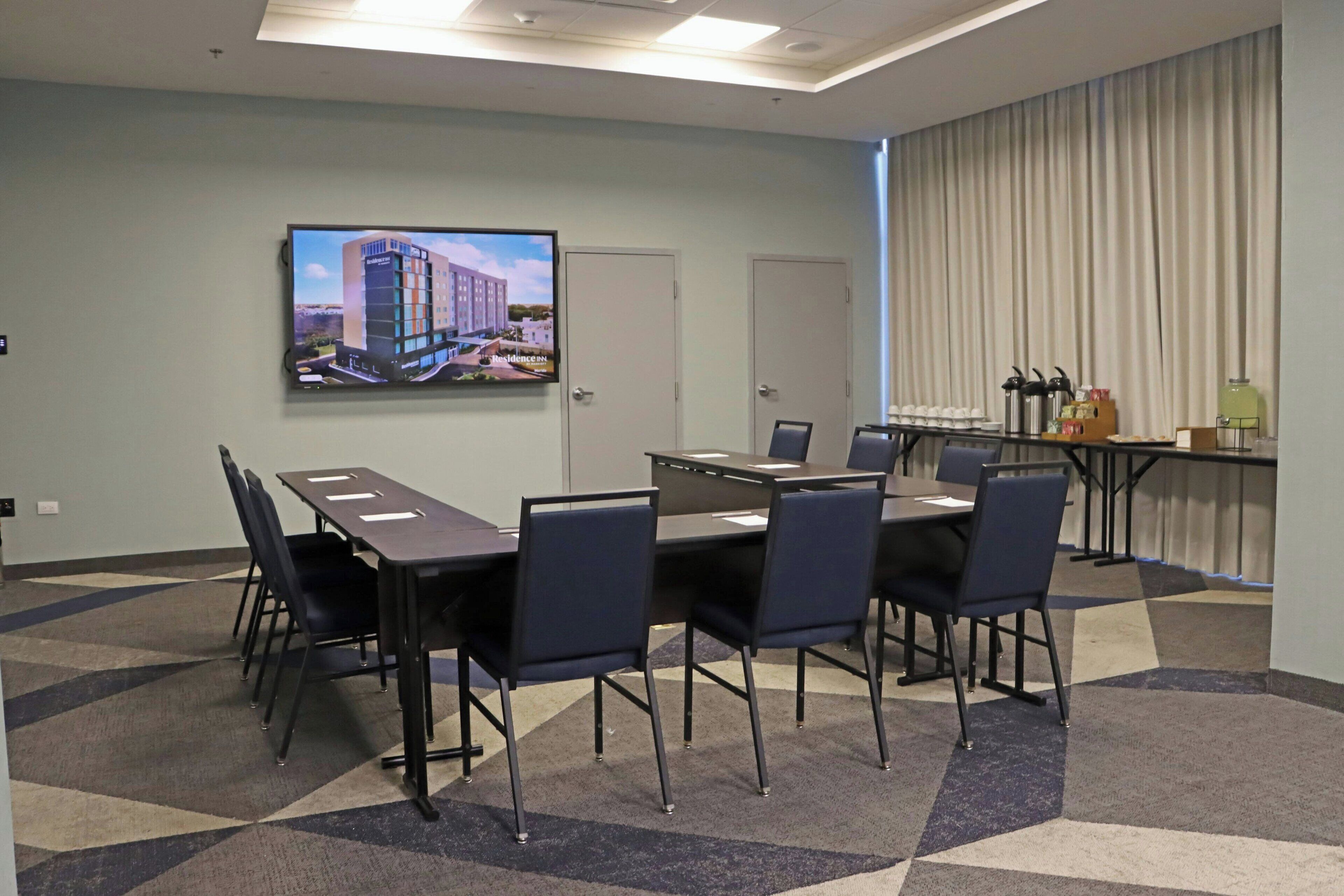 Un salón de conferencias con una pantalla grande mostrando una imagen de un edificio, una mesa larga con sillas y una zona de buffet con comida y bebidas.