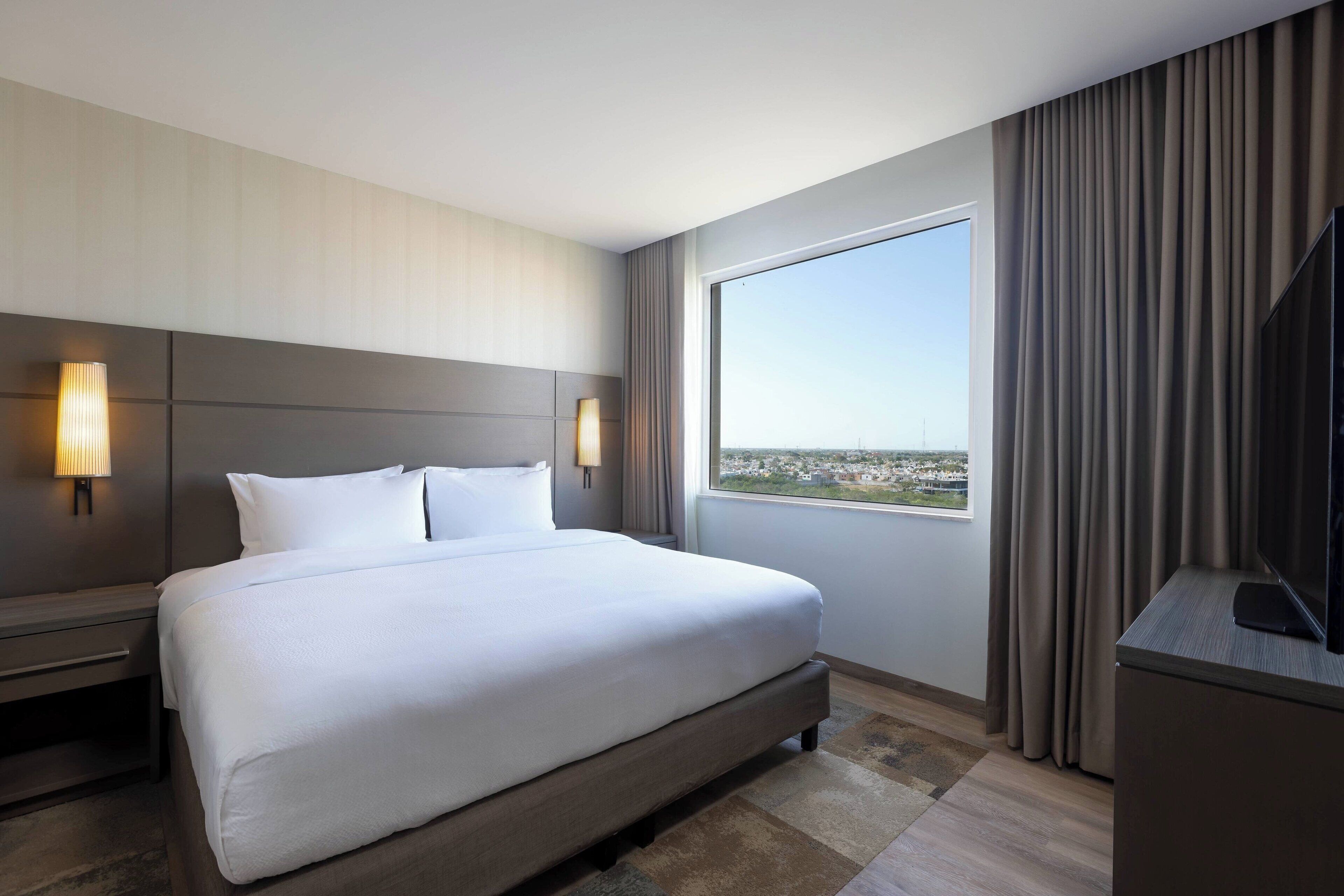 A hotel room with a large bed, two bedside lamps, a TV, a window with curtains, and a view of a cityscape.