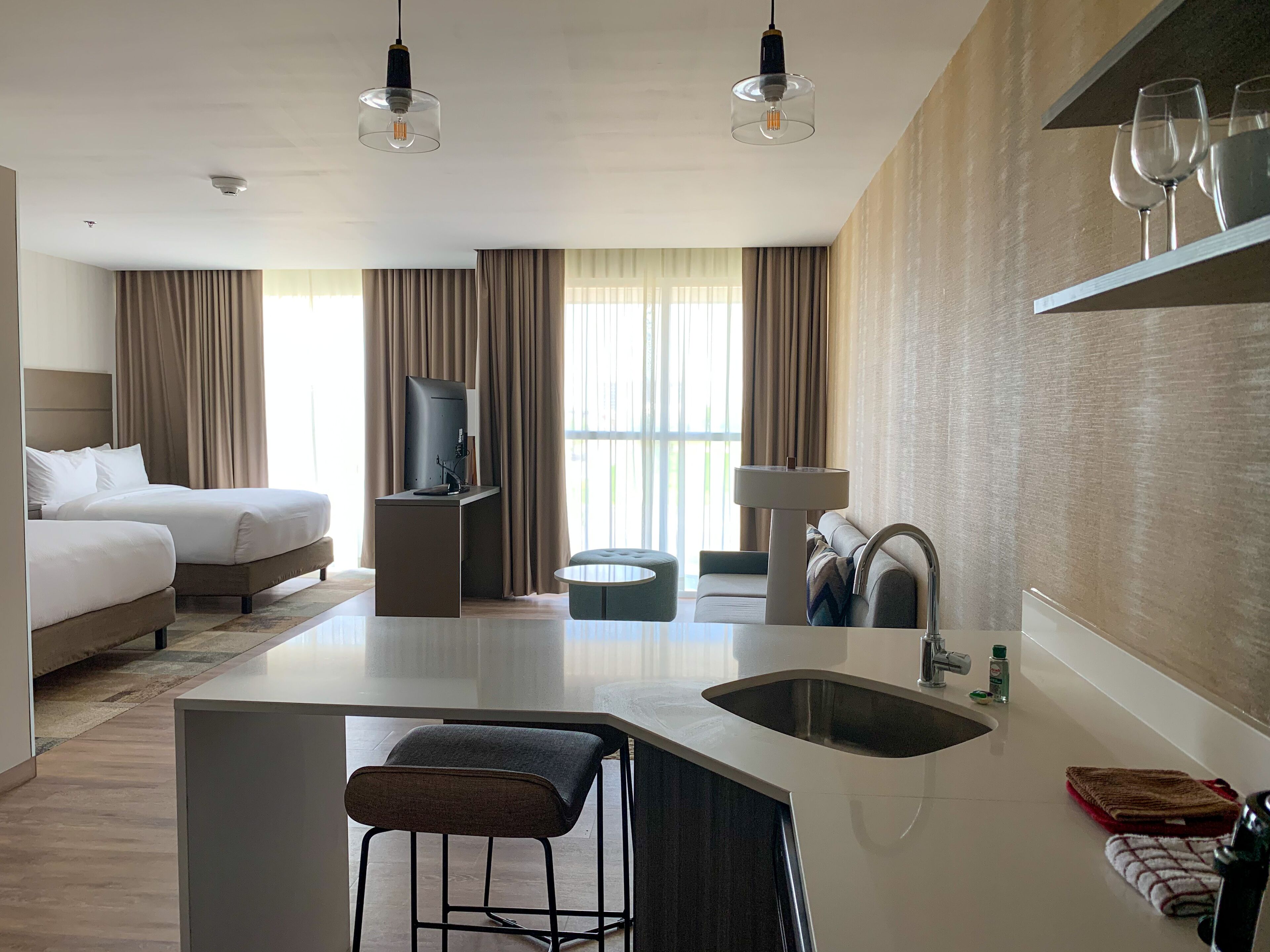 Habitación de hotel moderna con cama, escritorio con televisor, pequeña cocina y zona de estar con taburete de bar.
