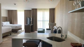 Habitación de hotel moderna con cama, escritorio con televisor, pequeña cocina y zona de estar con taburete de bar.