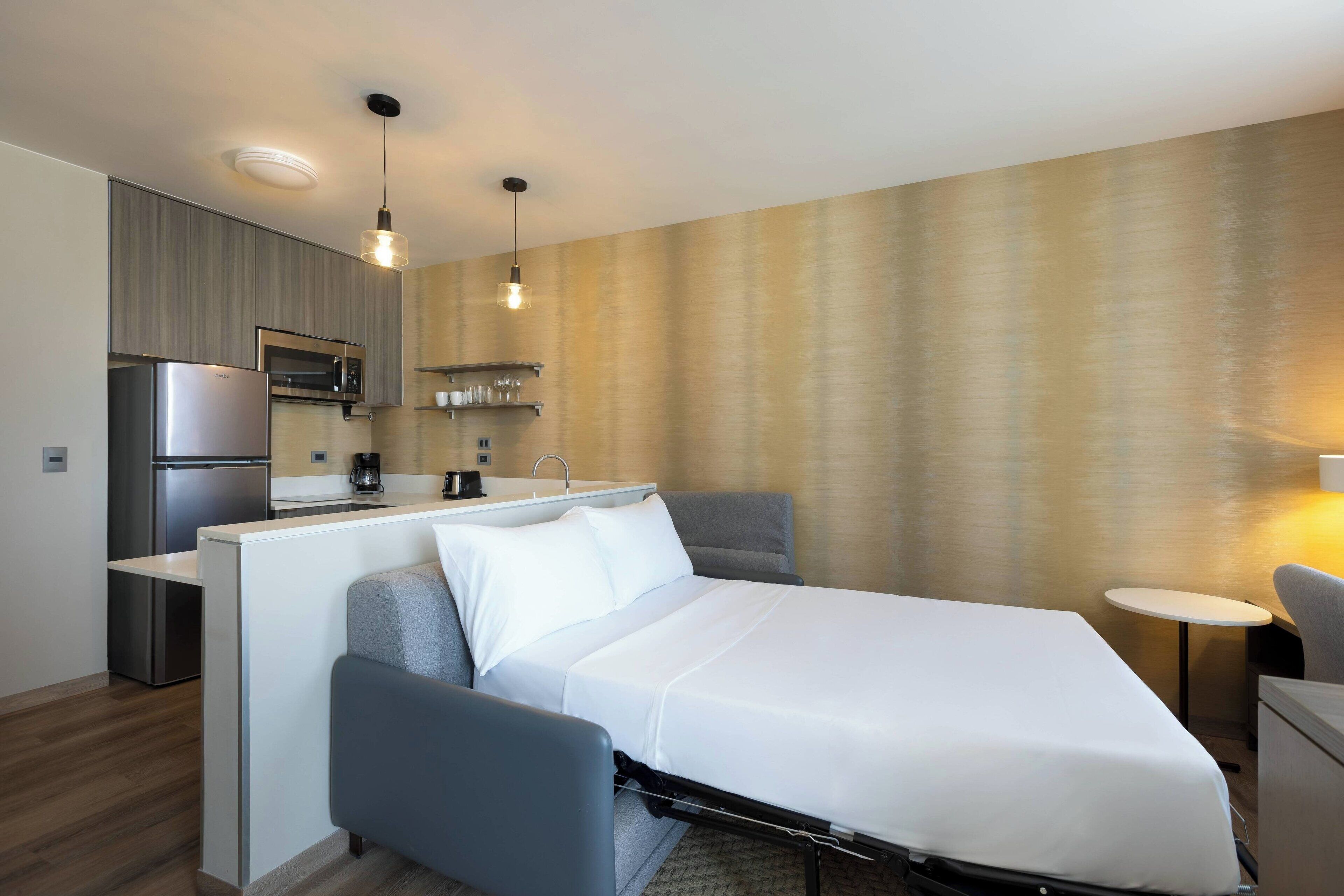 A compact hotel room with a bed, a small desk, a kitchenette, and a seating area.