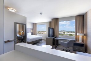 A modern hotel room with a bed, desk, chair, TV, and a view of the city.