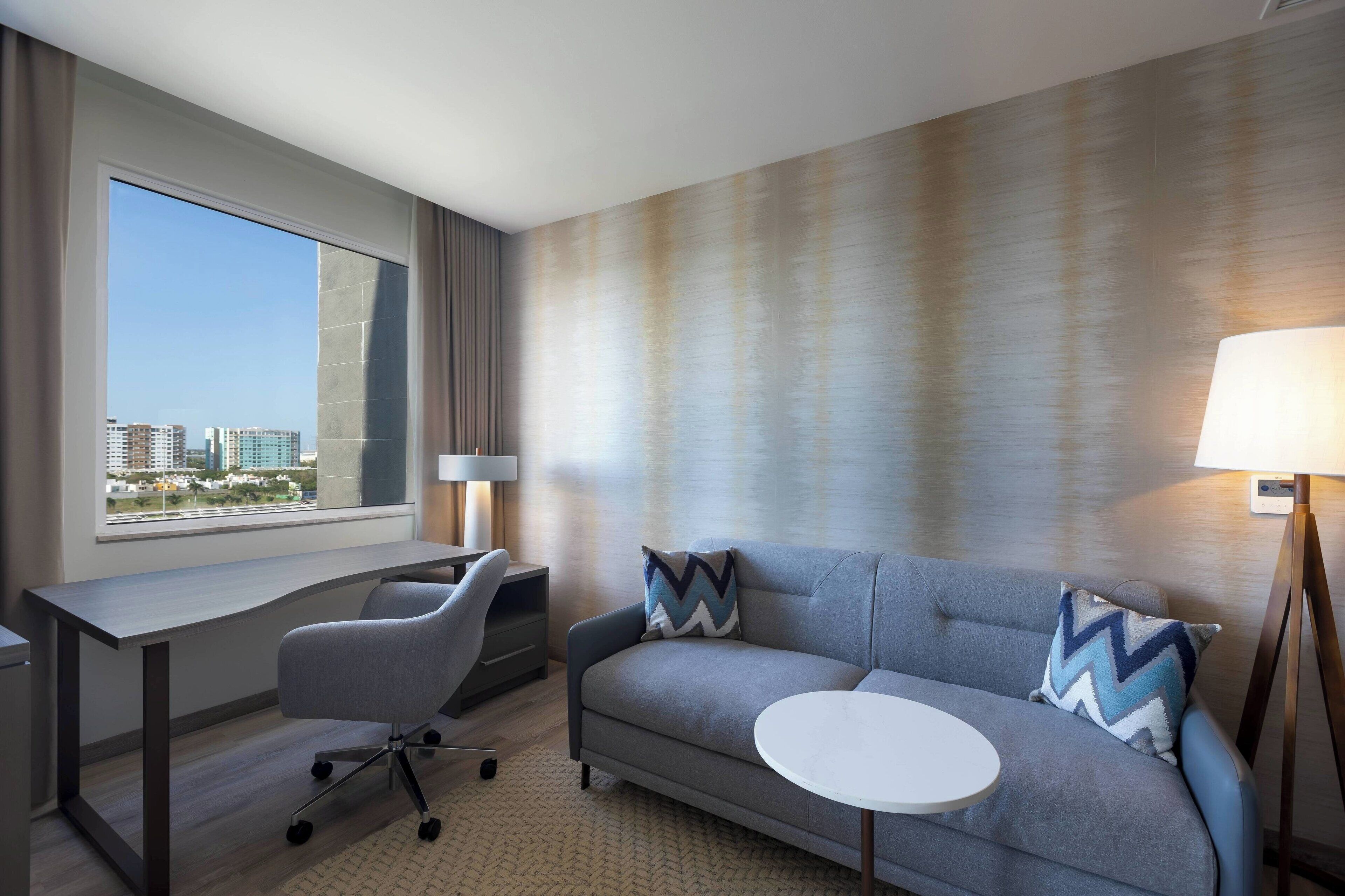 Habitación de hotel moderna con sofá gris, una pequeña mesa redonda, un escritorio y una silla. Hay un ventanal grande con vistas a la ciudad.