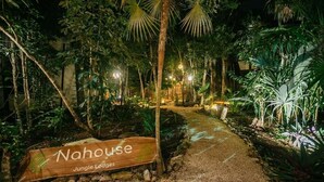 En sti der fører hen til et træskilt med teksten "Nahouse Jungle Lodges" omgivet af frodig vegetation og blidt oplyst.