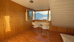 Ein Zimmer mit Holzwänden, Holzboden, einem Schreibtisch mit zwei Stühlen, einem Fenster mit Bergblick und einem Bett.