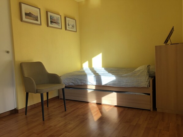 Una habitación pequeña y luminosa con una cama individual, una silla y un armario.