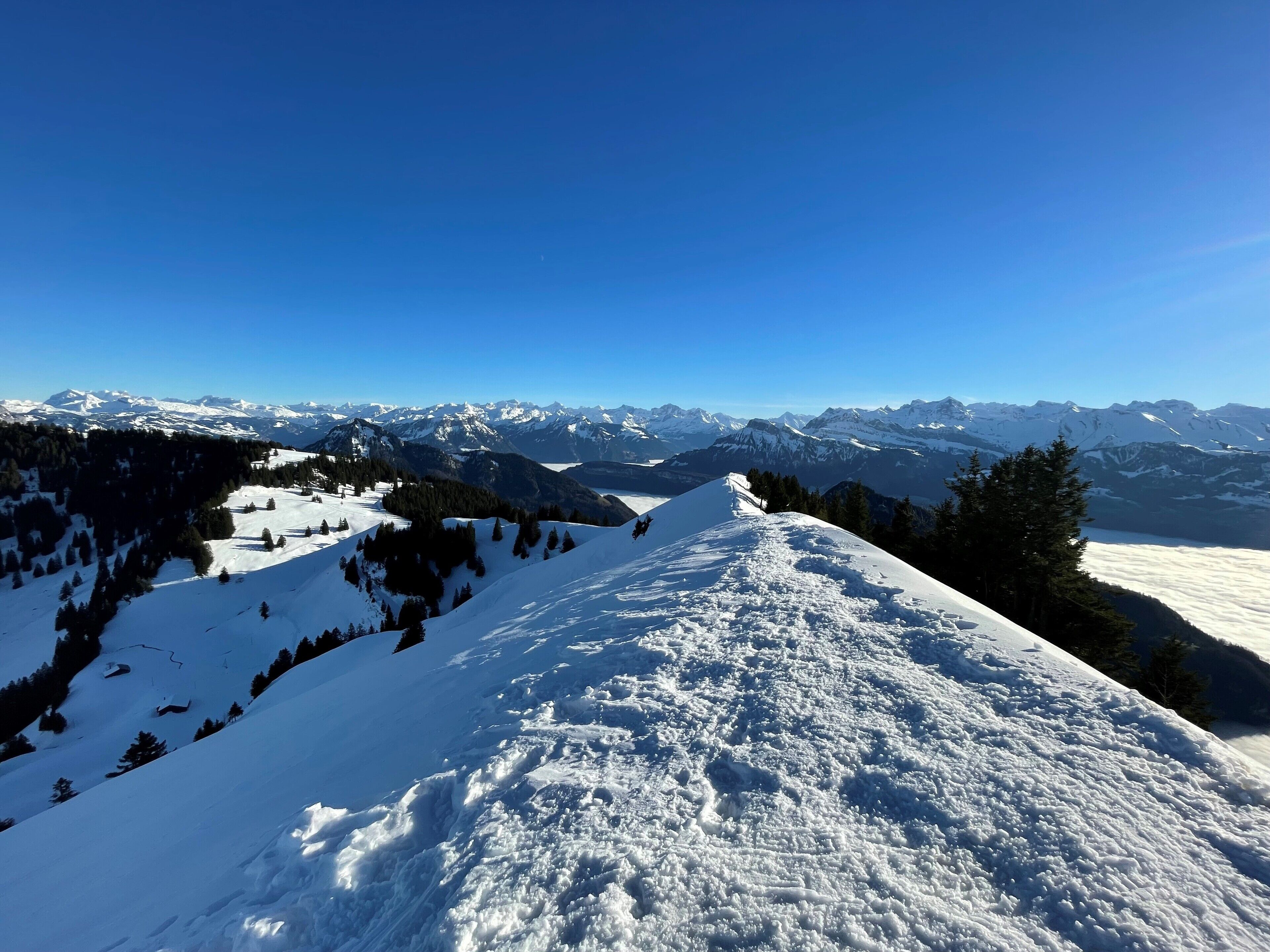Pendiente nevada de una montaña con una vista despejada de picos nevados a lo lejos, bajo un cielo azul.