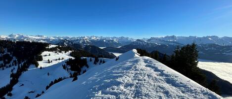 Pendiente nevada de una montaña con una vista despejada de picos nevados a lo lejos, bajo un cielo azul.