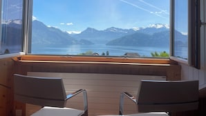 Ein Zimmer mit großem Fenster und Blick auf See und Berge. Zwei Stühle stehen im Raum.