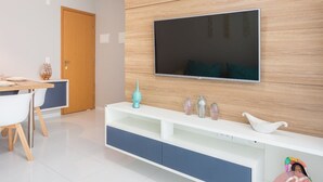 A modern living room with a flat-screen TV mounted on a wooden wall, a white entertainment unit with storage, a dining area with a wooden table and white chairs, and a door in the background. - Palm Village Acqua (Ipojuca)