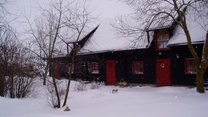Ein mit Schnee bedecktes Holzhaus mit roten Türen und Fenstern, umgeben von kahlen Bäumen und einer verschneiten Landschaft.