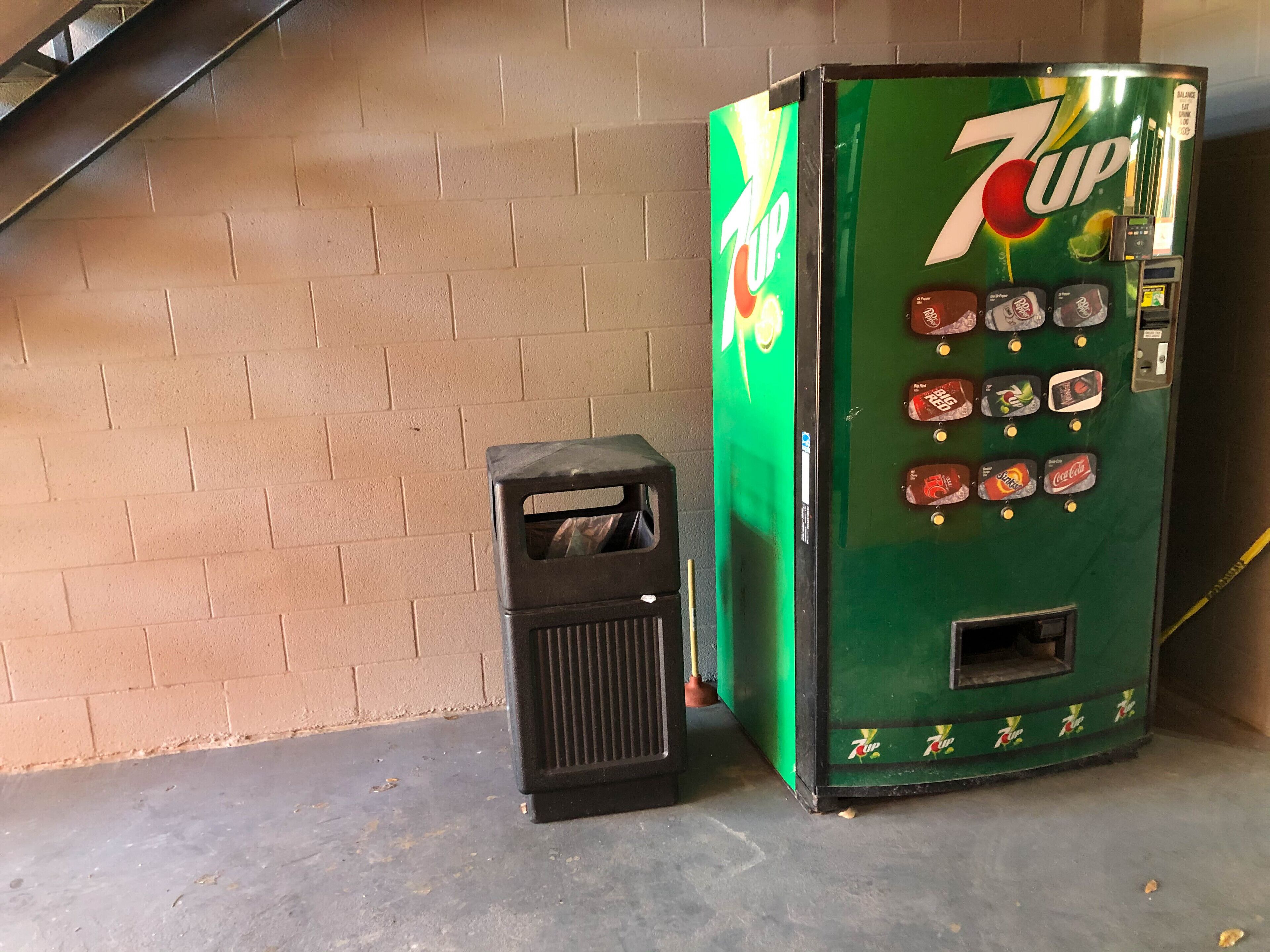 Een 7UP automaat met een muntgleuf en een prullenbak ernaast.