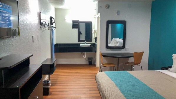 A hotel room with a bed, a desk, a chair, a small table, a sink, and a mirror.