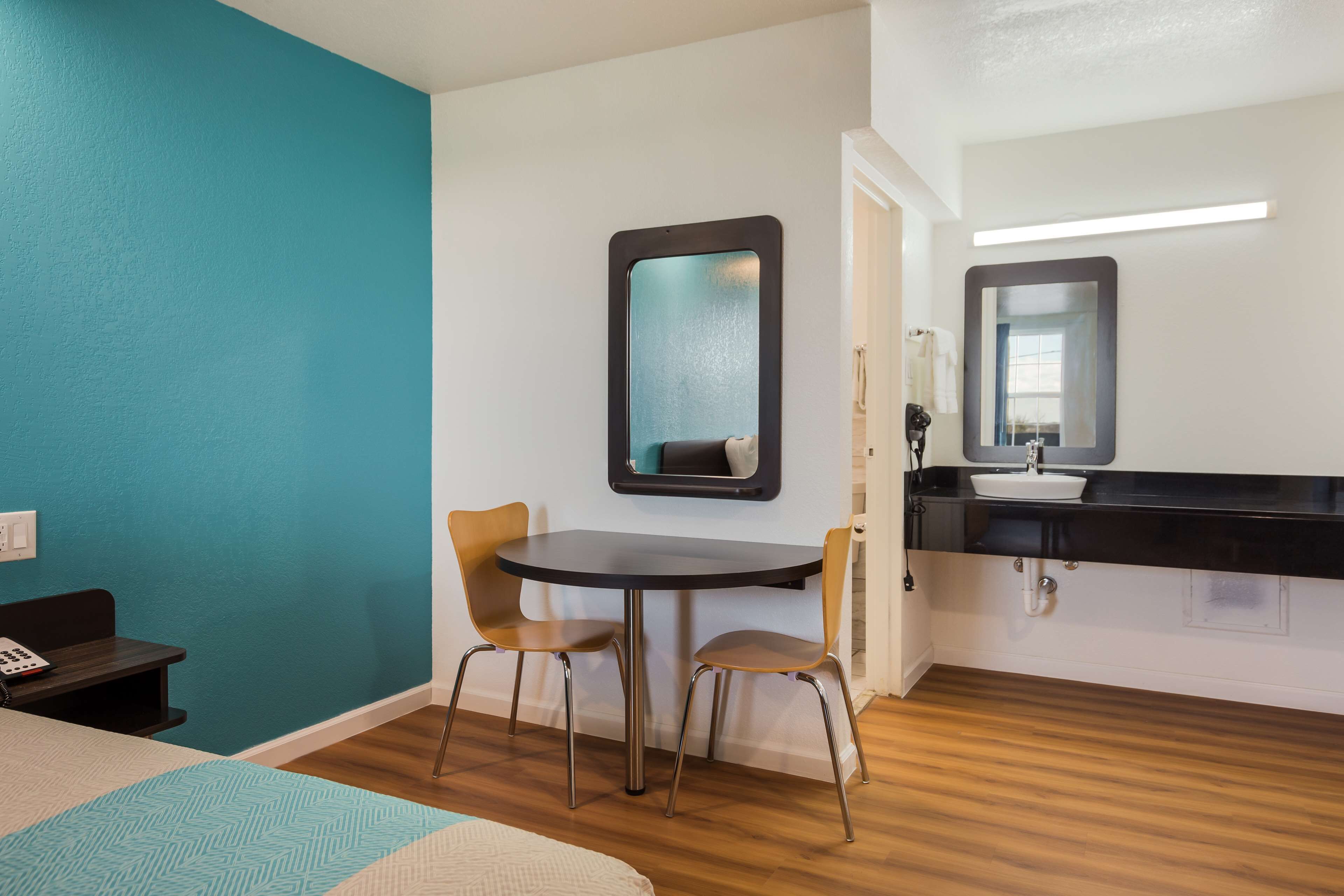 Habitación de hotel con paredes verde azulado, una mesa redonda con dos sillas, una cama con colcha estampada, una mesita de noche pequeña con un teléfono y un baño con lavabo y espejo.