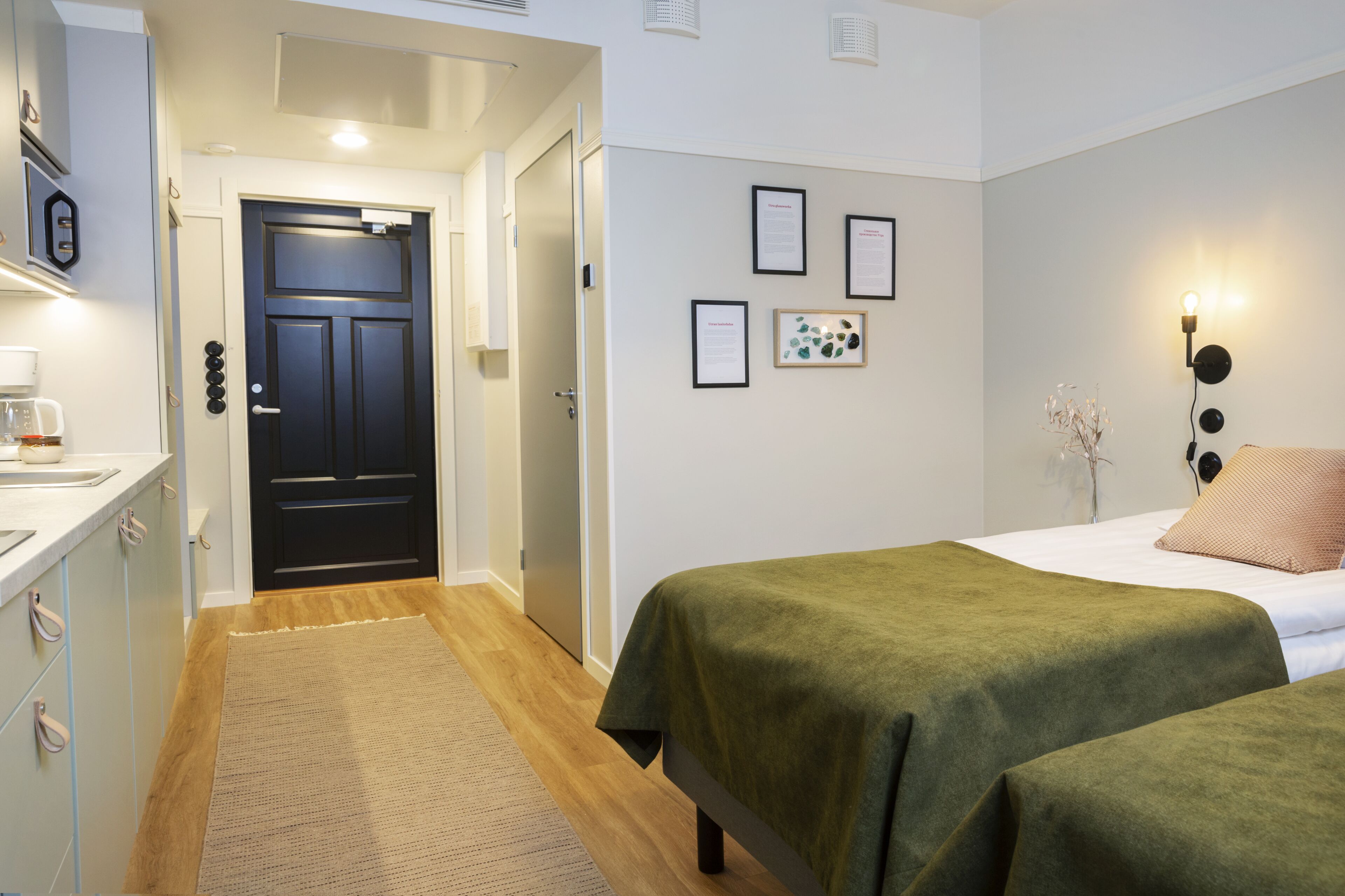 A hotel room with a bed, a desk with a lamp, a door, and framed certificates on the wall.