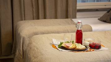 Una cama bien hecha con una bandeja de desayuno sobre un edredón color beige, que incluye una botella de bebida roja, un vaso de agua y varios alimentos.