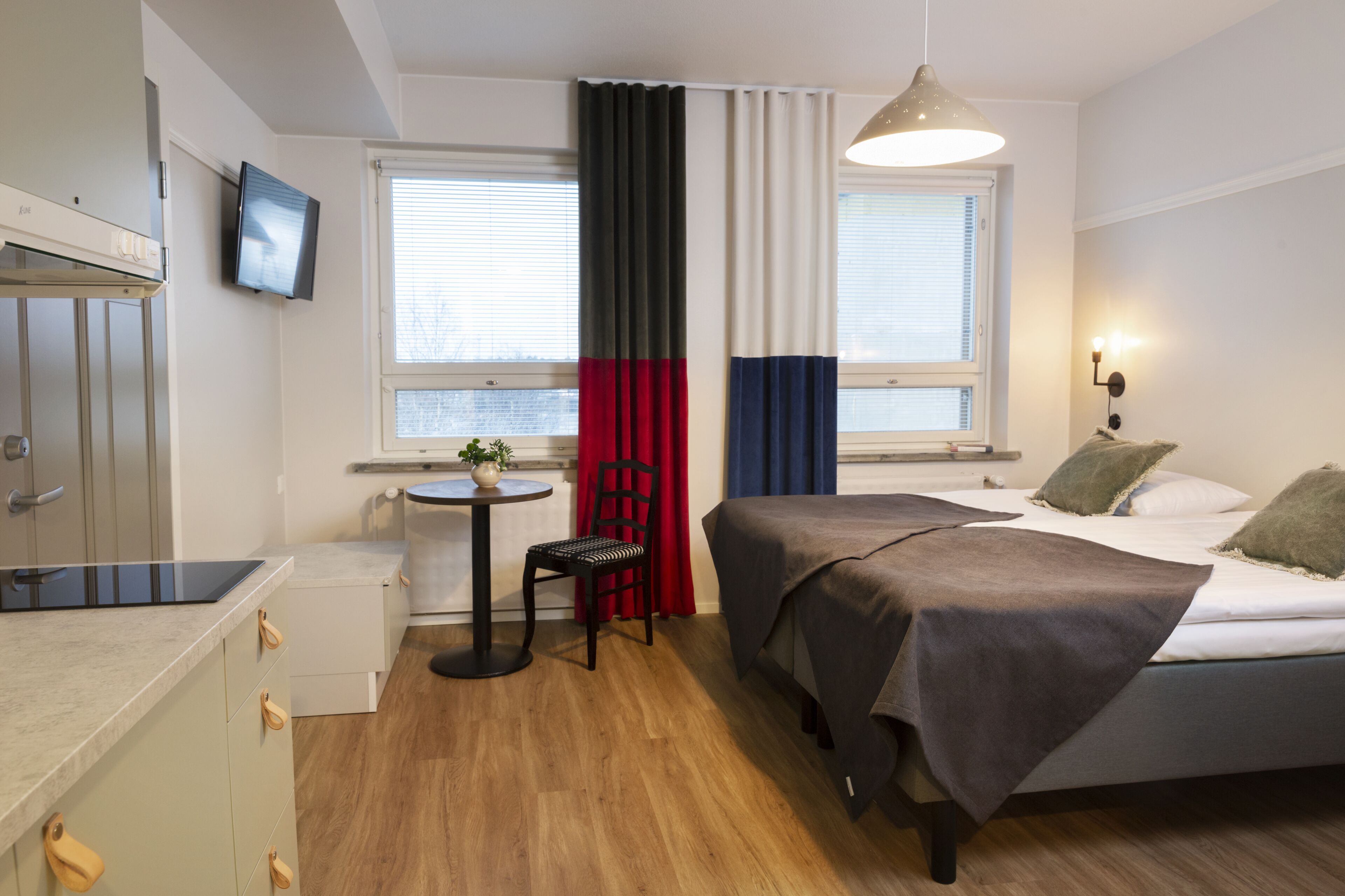 A compact hotel room with a bed, a small table with a chair, a TV mounted on the wall, and a window with curtains.