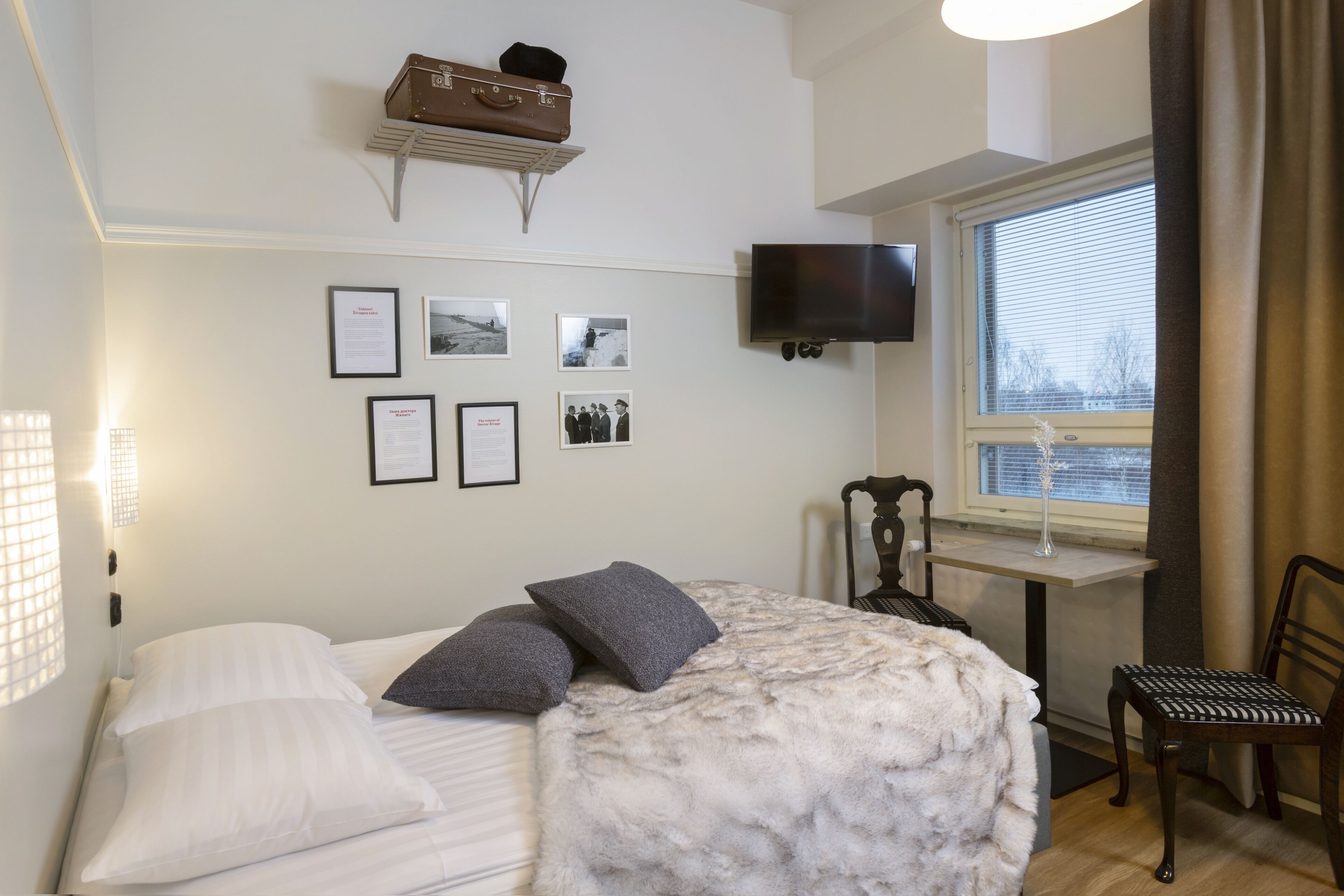 Un dormitorio con cama, almohadas, televisor montado, cuadros en la pared, ventana con persianas y una silla.