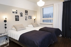 Une chambre avec deux lits, une table de chevet, une lampe fixée au mur et une fenêtre avec des stores.