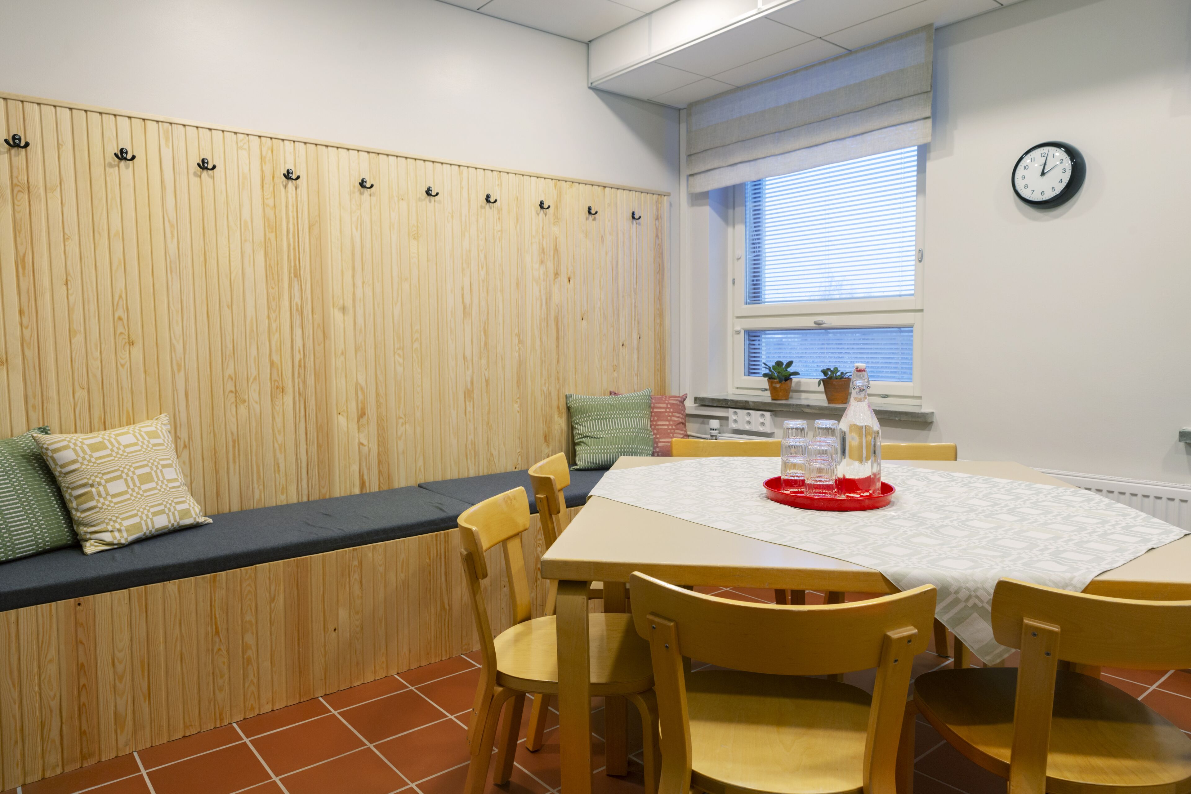 A dining area with a wooden bench, a table with chairs, a clock on the wall, and a window with blinds.