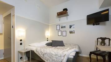 A bedroom with a bed, bedside table, lamp, wall-mounted TV, framed pictures, and a suitcase on a shelf.