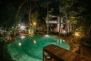 A nighttime pool area with clear water, surrounded by trees and a wooden deck with seating.
