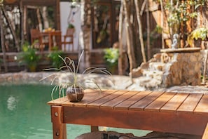 A wooden table by a pool with a potted plant, surrounded by natural stone and greenery.