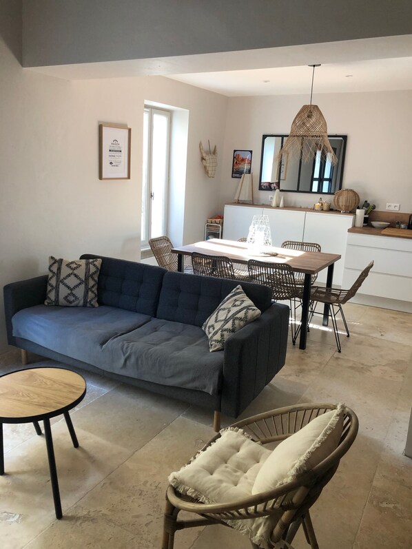 Un salon moderne avec un canapé bleu foncé, une table basse ronde en bois et un fauteuil en osier. On aperçoit à l’arrière-plan un coin repas avec une table et des chaises en bois.
