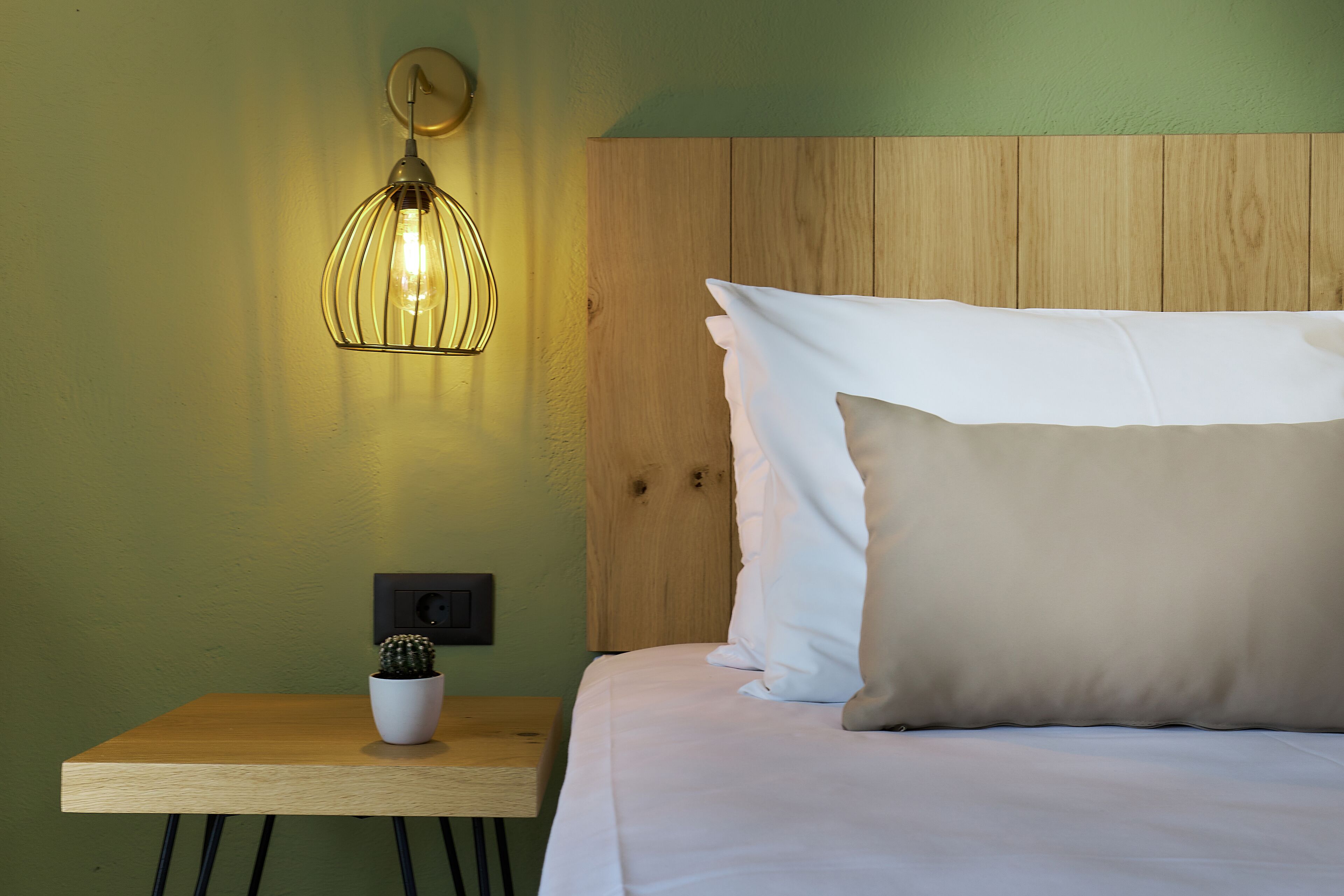 Een opgemaakt bed met witte en beige kussens, een kleine cactus in een pot op een nachtkastje van hout, en een wandlamp.