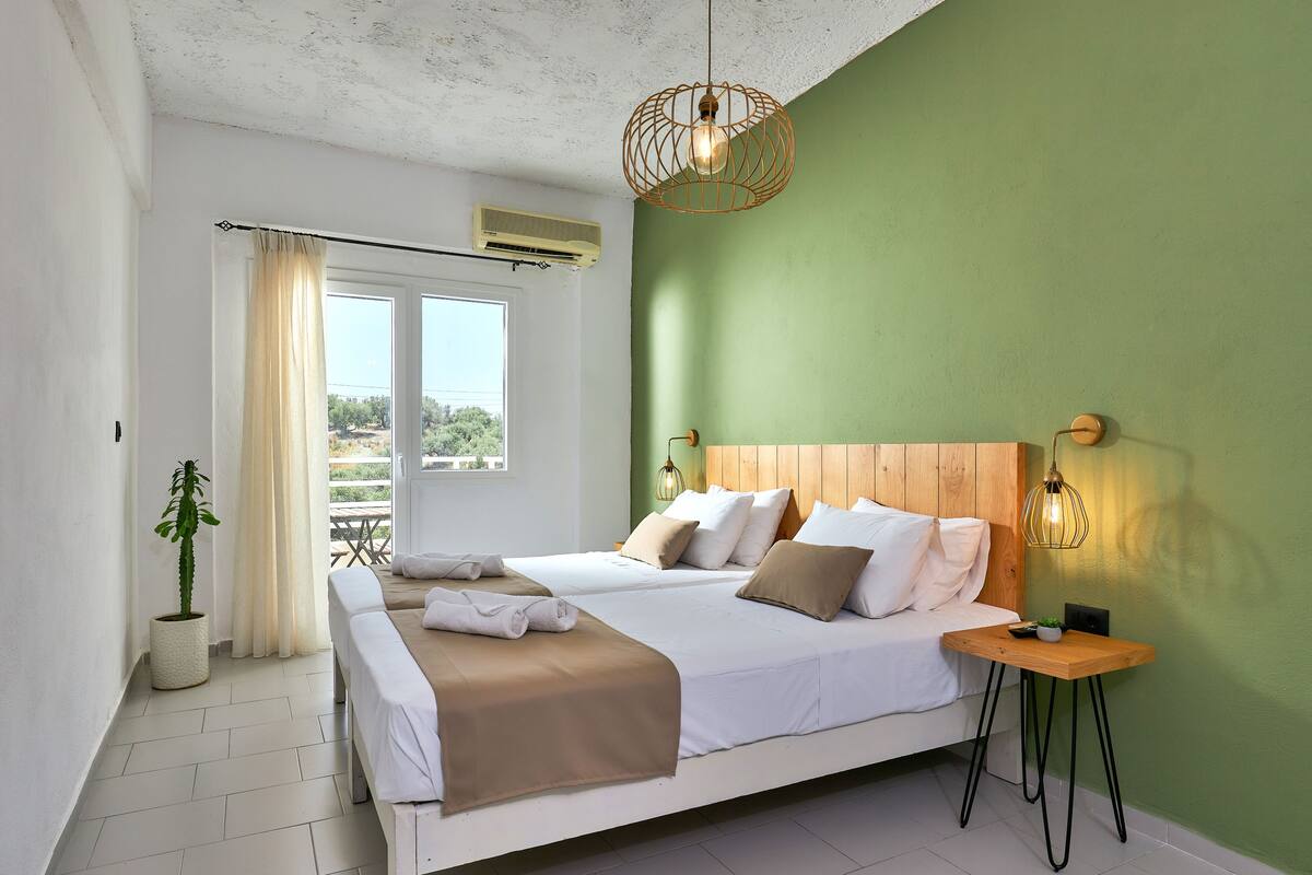 A double bed with white and brown bedding, a bedside table with a lamp, and a view of the outdoors through a window with curtains.