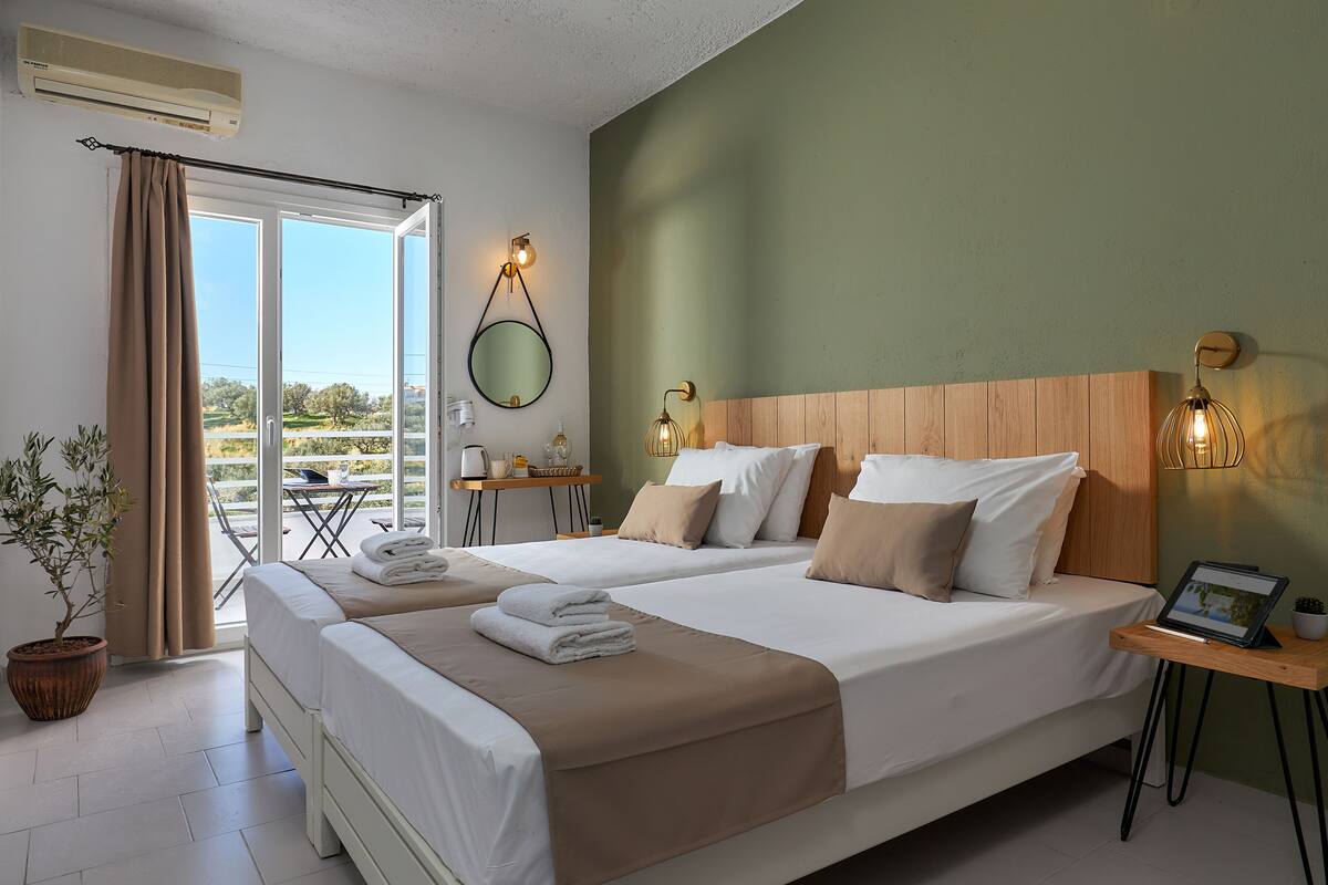 A hotel room with two beds, a wooden headboard, a small table with a laptop, and a view of the outdoors.