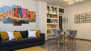 A modern living room with a grey sofa, a yellow rug, a dining table with chairs, and a bookshelf with books and decorative items.