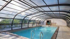 Una piscina con techo de vidrio, rodeada por una terraza de madera y una barandilla de metal.