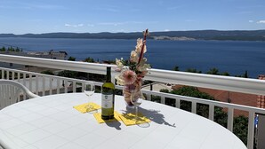 Et bord med en vinflaske, et glas og blomsterudsmykning på en balkon med udsigt over vand og fjerne bakker.