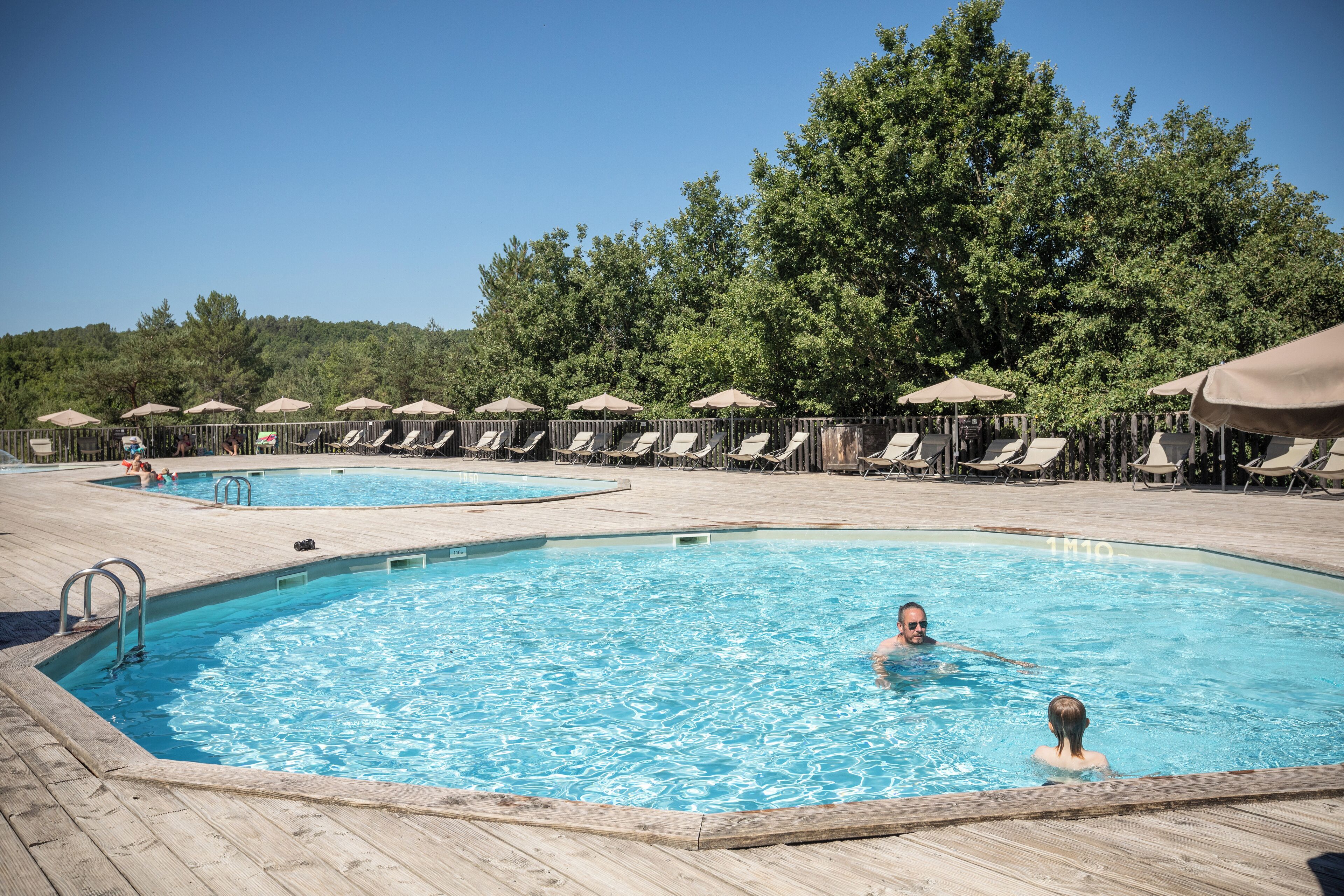 A pool area with multiple lounge chairs and umbrellas, surrounded by trees.