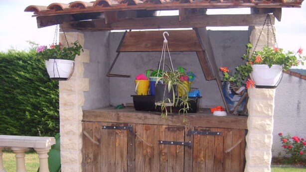 A rustic outdoor kitchen with a wooden structure, hanging planters with flowers, and a stone wall in the background.