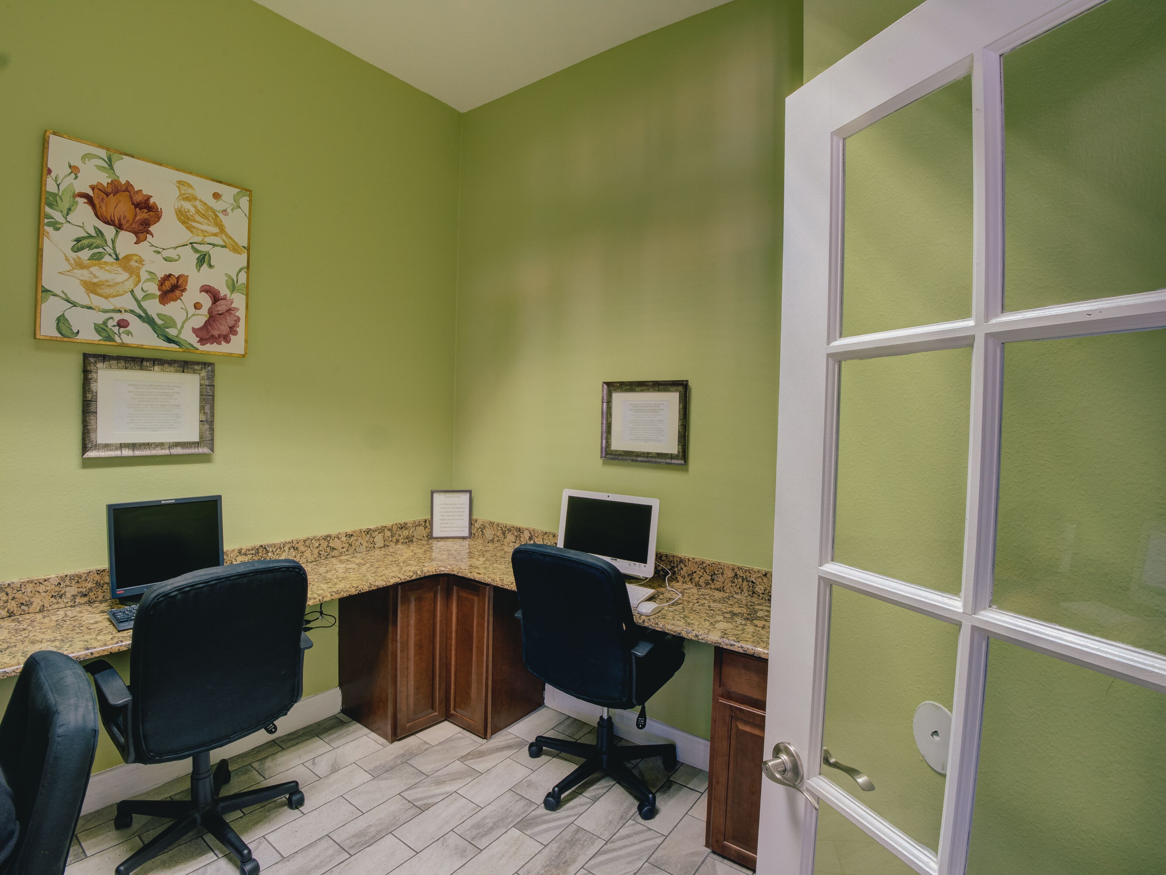 A room with green walls, a wooden desk, two black office chairs, two computer monitors, and a door with frosted glass panels.