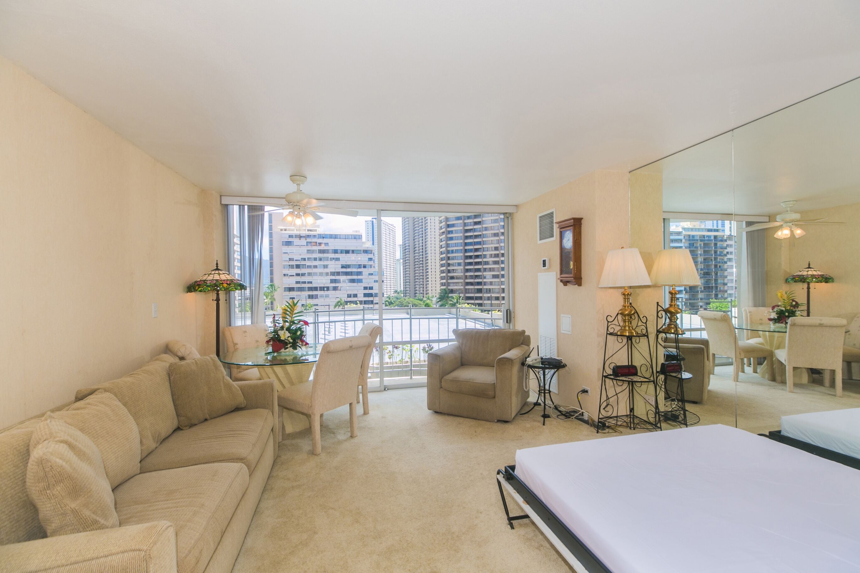 A hotel room with a large bed, a sofa, a dining table with chairs, and a balcony with a view of tall buildings.