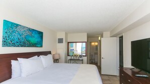 Quarto de hotel com cama grande, televisão, escrivaninha com candeeiro e um quadro na parede.
