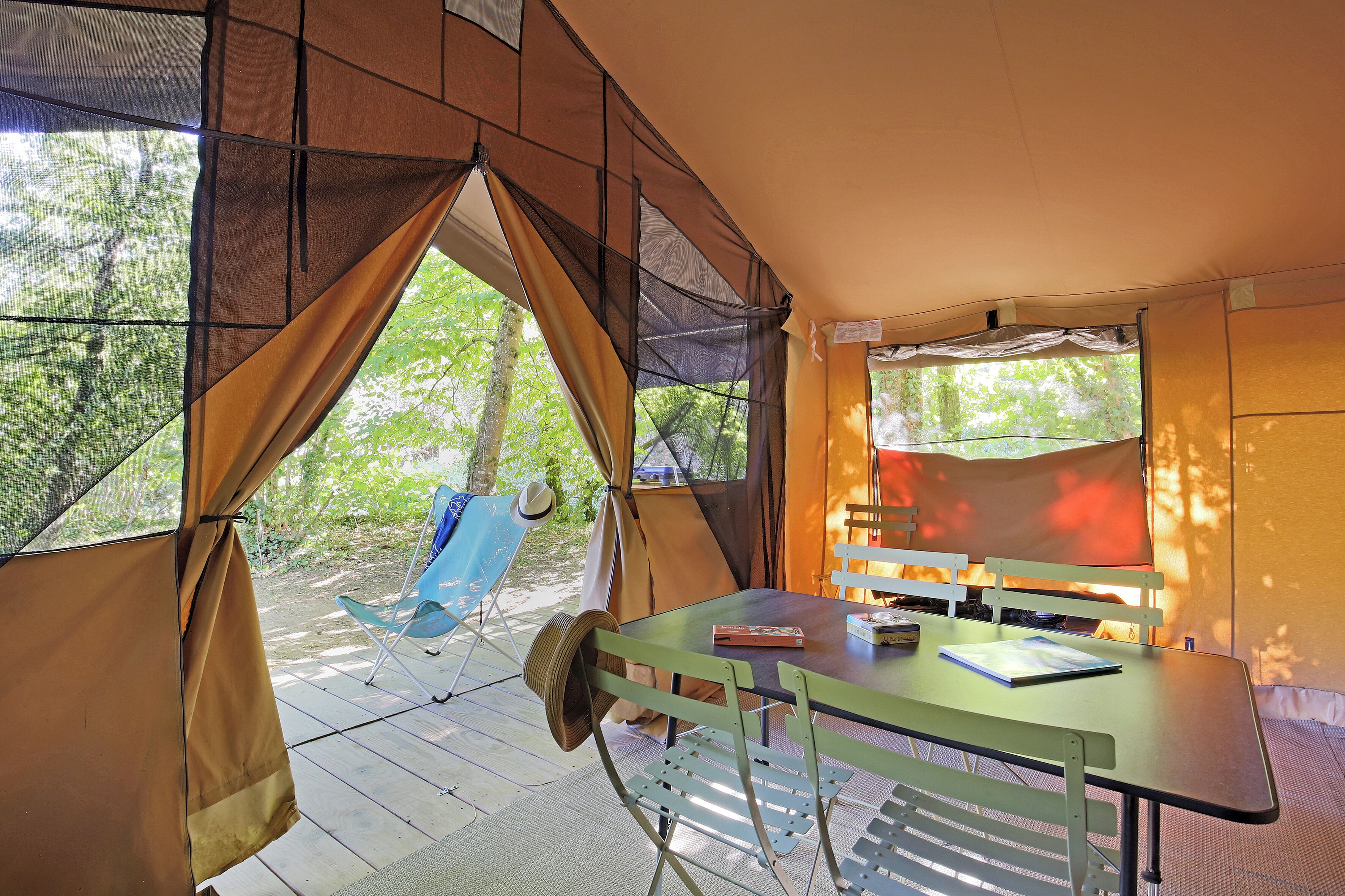 Une tente avec une table et des chaises, un chapeau posé sur une chaise, et une vue sur les arbres à l’extérieur.