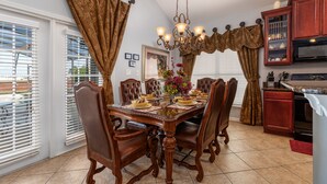A dining room with a wooden table set for six, surrounded by six chairs. There is a chandelier above the table, and a kitchen area is visible to the right.