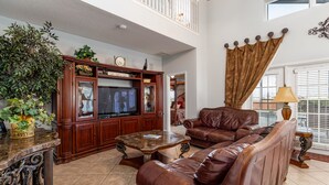 A spacious living room with a brown leather sofa, a wooden entertainment center, and a large window with blinds.