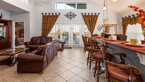 A spacious living room with a high ceiling, a dining area with wooden chairs and a bar, and a view of the outdoors through large windows.